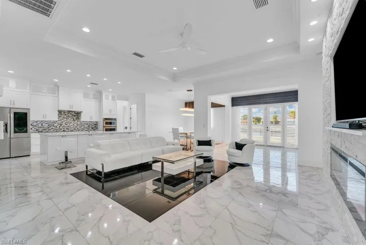 Open-concept living space featuring high ceilings with recessed lighting, an integrated kitchen with stainless steel appliances and white cabinetry, a built-in fireplace with a stone surround, and large French doors with interior shutters
