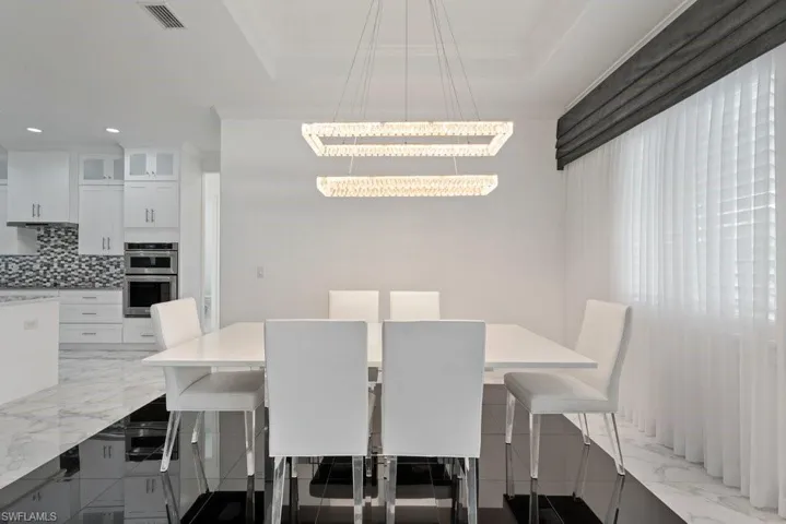Dining area featuring a modern chandelier, white walls, and a large window with sheer white curtains and a dark valance