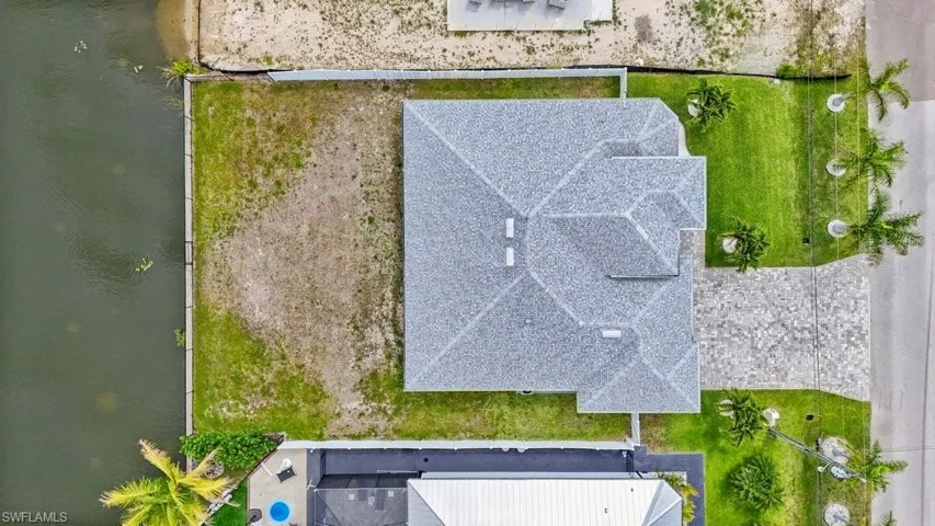 Waterfront property featuring a gray shingle roof, green lawn, a concrete driveway, and palm trees