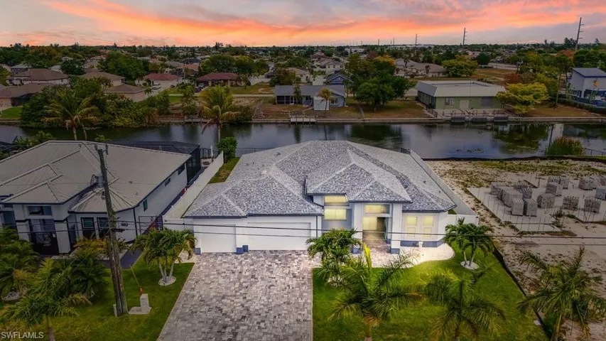 Waterfront property featuring a paver driveway, mature landscaping, multi-peak roofline, and an integrated two-car garage