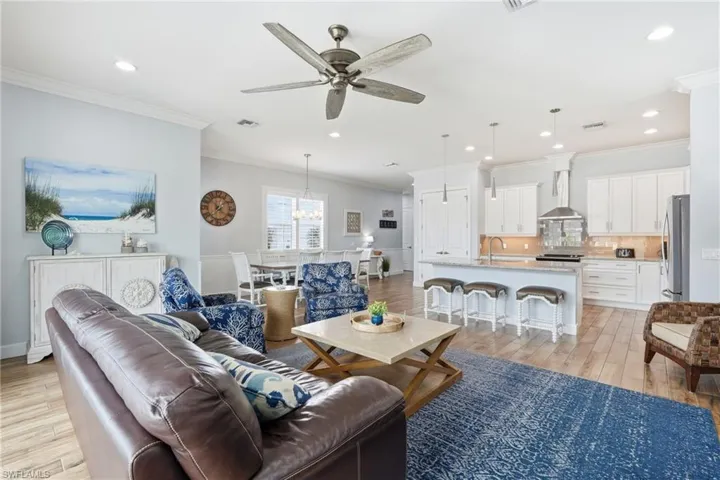 Open-concept living area with wood-finish flooring, recessed lighting, a ceiling fan, and white kitchen cabinetry with a central island - Virtually Edited Image
