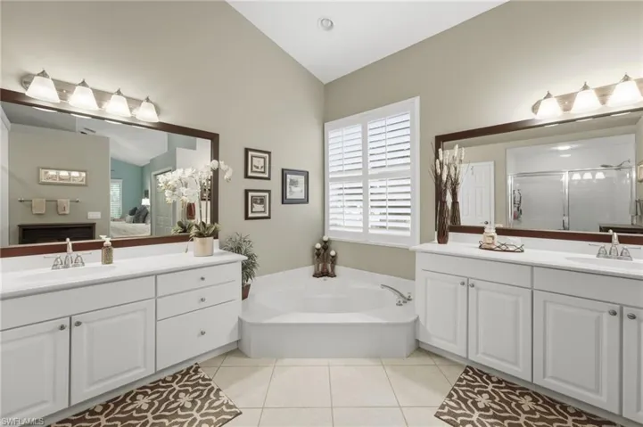 Spacious bathroom featuring a built-in corner soaking tub, dual vanities with white cabinetry, large framed mirrors, and plantation shutters