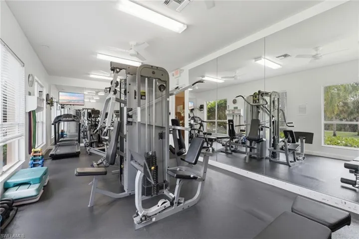 Spacious fitness room featuring a full wall of mirrors, multiple exercise machines, rubber flooring, and ample natural light from several windows