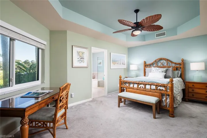 Carpeted bedroom with visible vents, a tray ceiling, baseboards, a ceiling fan, and ensuite bath