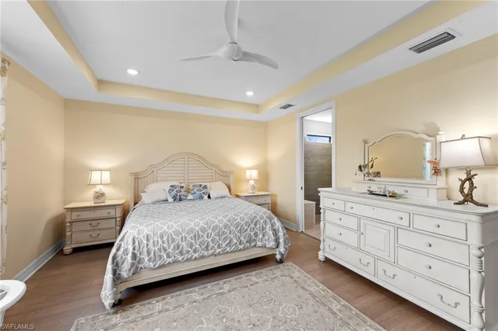 Bedroom with light wood finished floors, a raised ceiling, ensuite bath, ceiling fan, and recessed lighting