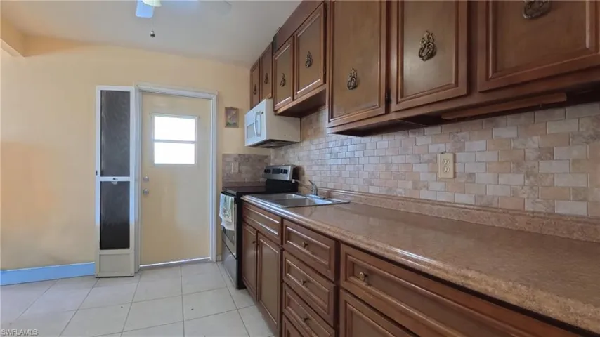 Kitchen with electric stove, tasteful backsplash, white microwave, light tile patterned floors, and brown cabinetry