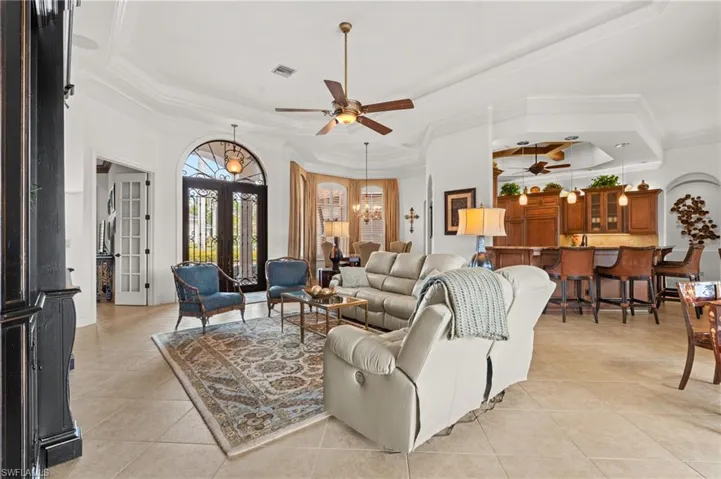 Tiled living room featuring ceiling fan with notable chandelier, a tray ceiling, ornamental molding, and french doors