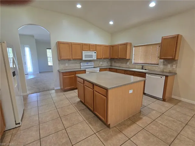 Kitchen featuring decorative backsplash, arched walkways, white appliances, a center island, and brown cabinetry