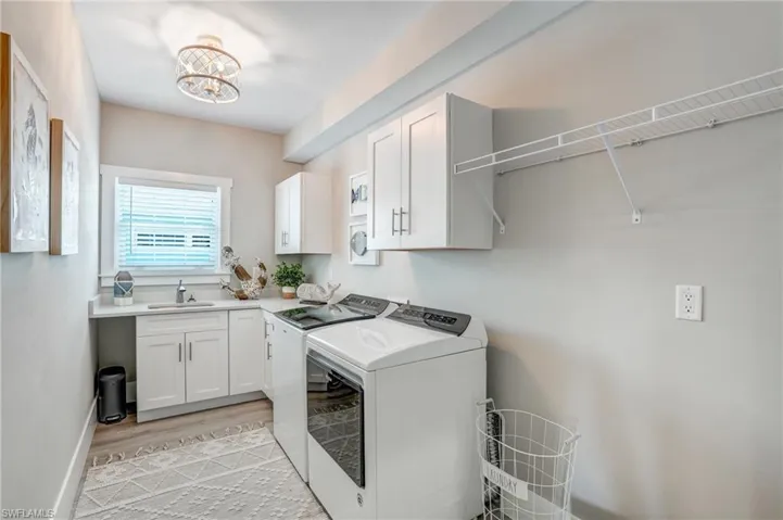Laundry room featuring washing machine and dryer, light wood-style floors, cabinet space, and a chandelier - Virtually Edited Image
