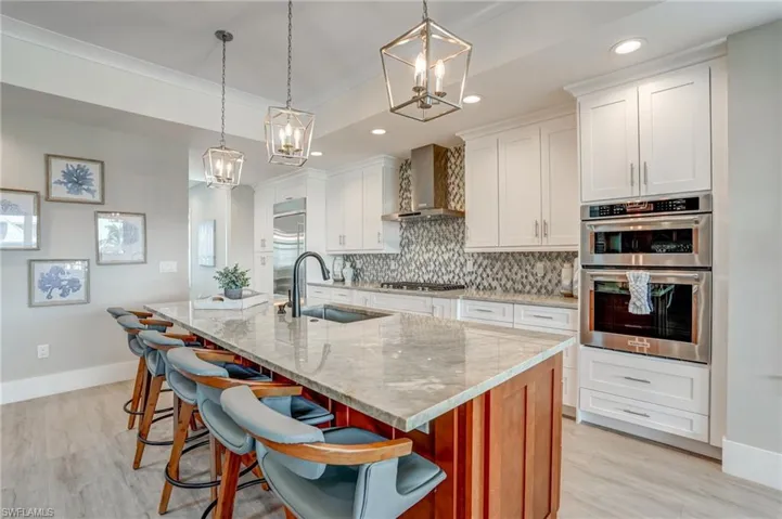 Kitchen featuring a kitchen island with sink, backsplash, light stone counters, hanging light fixtures, and white cabinets - Virtually Edited Image