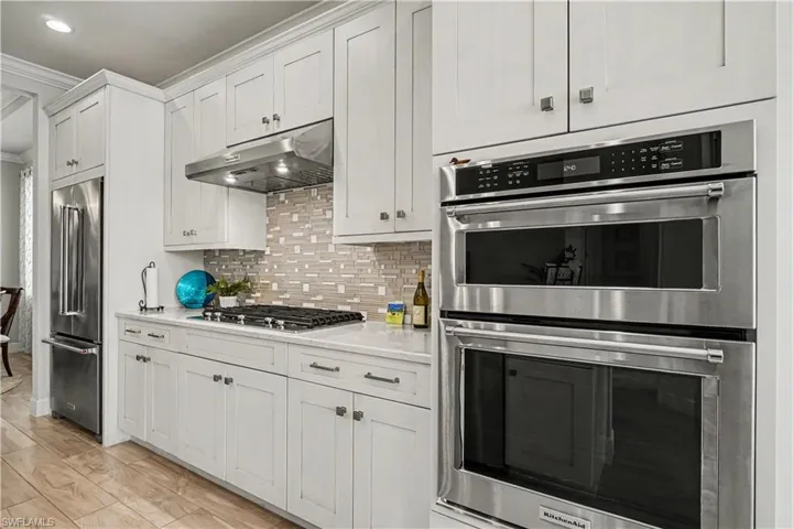 White shaker-style cabinetry with brushed nickel hardware, stainless steel Kitchen Aide appliances, mosaic tile backsplash, and tile flooring