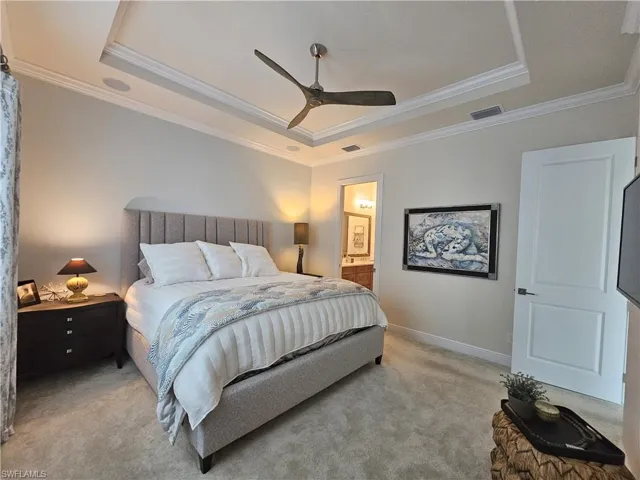 Carpeted bedroom with a tray ceiling, crown molding, ceiling fan, and connected bathroom