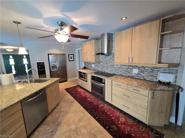 Kitchen featuring light brown cabinets, stainless steel appliances, wall chimney range hood, tasteful backsplash, and a ceiling fan