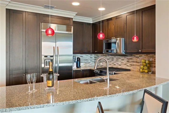 Kitchen featuring stainless steel appliances, light stone countertops, dark brown cabinets, crown molding, and hanging light fixtures