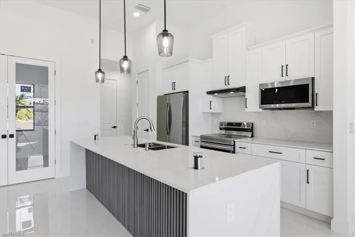 Kitchen with a kitchen island with sink, stainless steel appliances, light stone counters, white cabinetry, and hanging light fixtures