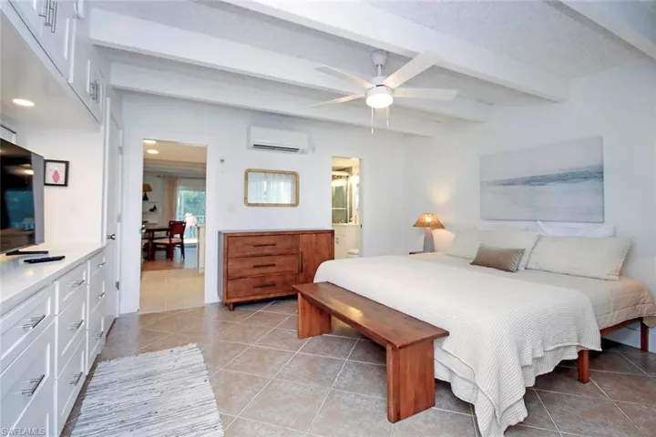 Bedroom with light tile patterned flooring, beamed ceiling, ensuite bath, ceiling fan, and a wall unit AC