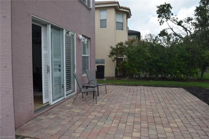 Paved patio featuring multi-toned pavers, direct access via sliding glass doors with interior shutters, and a surrounding privacy hedge