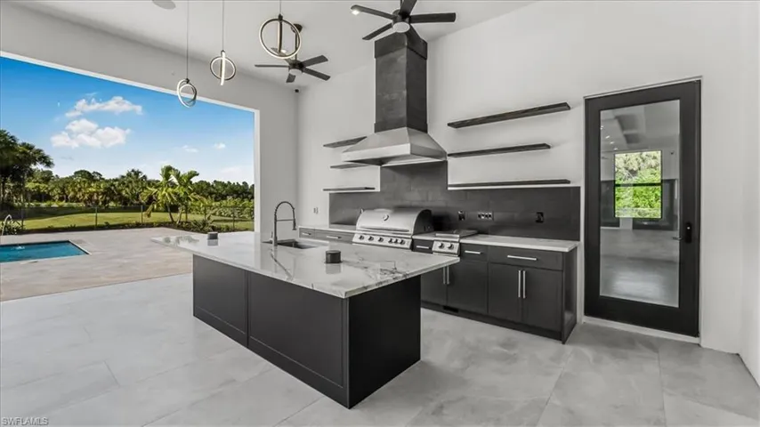 Kitchen featuring dark cabinets, decorative light fixtures, a center island with sink, decorative backsplash, and light stone countertops