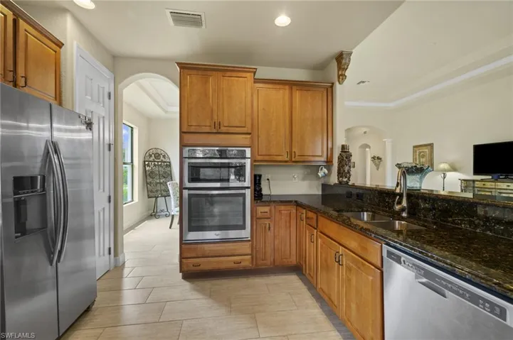 Kitchen with arched walkways, dark stone countertops, stainless steel appliances, brown cabinetry, and recessed lighting