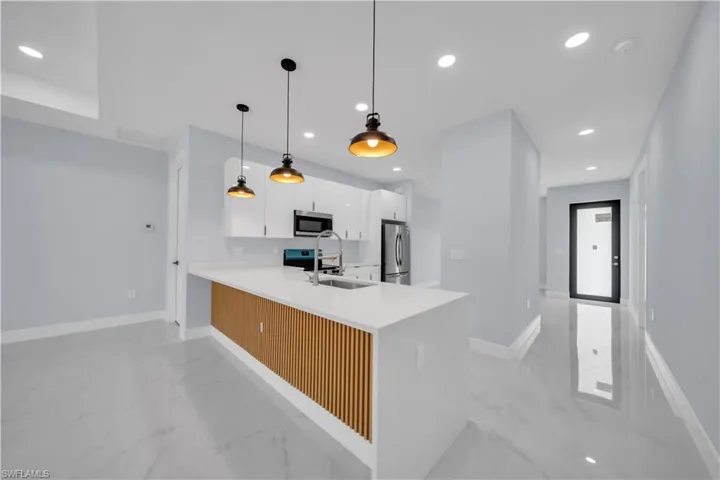 Open-concept kitchen featuring an island with a white countertop and wood-slat detailing, white flat-panel cabinetry, stainless steel appliances, and industrial-style pendant lighting