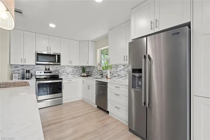 Modern kitchen featuring white cabinetry with brushed nickel hardware, stainless steel appliances, a multi-tone tile backsplash, light wood-finish flooring, and a window above the sink
