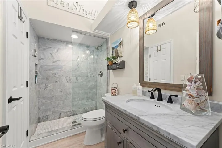 Bathroom featuring a glass-enclosed shower with tiled walls and a mosaic floor, a single vanity with a stone countertop, and a large framed mirror with two woven pendant lights