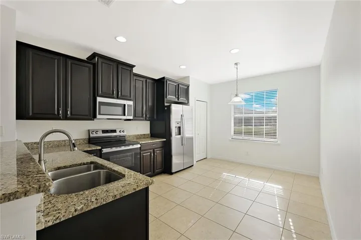 Kitchen featuring dark cabinetry, appliances with stainless steel finishes, light stone counters, decorative light fixtures, and light tile patterned floors