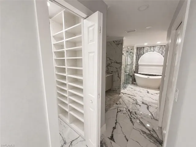 Built-in shelving storage, bi-fold closet doors, freestanding soaking tub, glass-enclosed shower, and large format marble-look tile flooring
