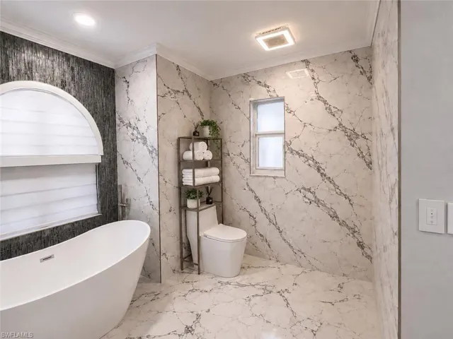 Freestanding soaking tub, floor-to-ceiling marble-look tile, textured accent wall, recessed lighting, and a contemporary toilet