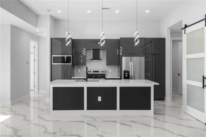 Kitchen featuring wall chimney exhaust hood, stainless steel appliances, pendant lighting, a barn door, and an island with sink