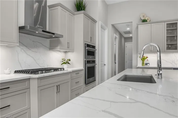 Kitchen with gray cabinetry, light stone countertops, sink, stainless steel appliances, and wall chimney exhaust hood.
