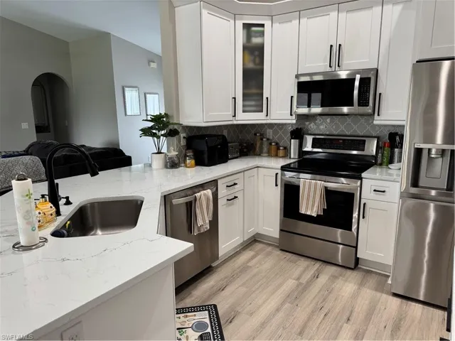 Modern kitchen featuring white shaker-style cabinetry, stainless steel appliances, a deep single-basin sink with a matte black faucet, light-toned wood-finish flooring, and a decorative tile backsplash