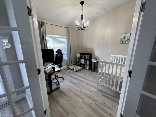French doors open to a versatile room featuring vaulted ceilings, wood-finish flooring, a contemporary chandelier, and a large window with blinds