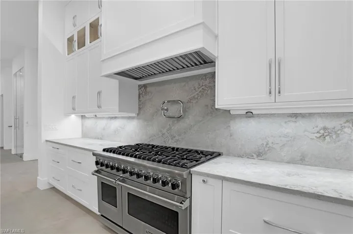 Kitchen with double oven range, light stone counters, decorative backsplash, white cabinetry, and custom exhaust hood