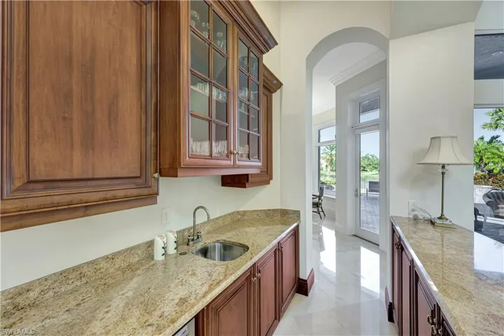 Bar in living room, glass insert cabinets, crown molding, light stone countertops, and arched walkways