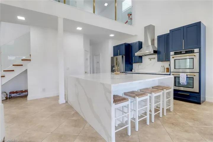 Kitchen featuring light tile patterned flooring, a high ceiling, wall chimney exhaust hood, appliances with stainless steel finishes, and blue cabinetry