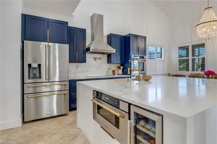 Kitchen featuring wall chimney range hood, beverage cooler, blue cabinetry, stainless steel appliances, and a center island with sink