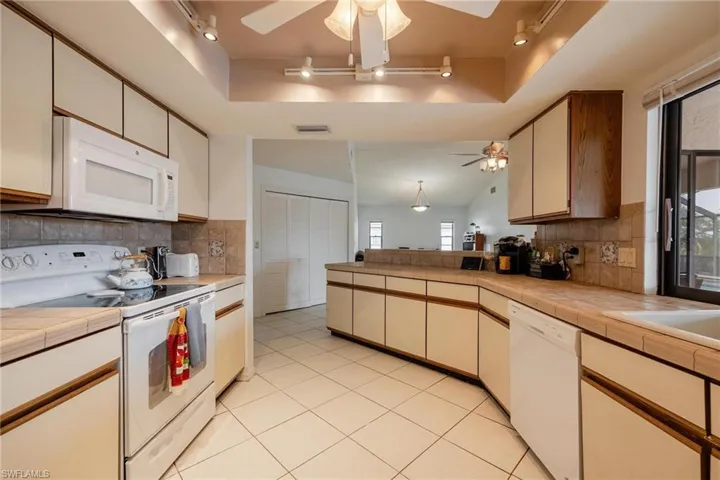 Two tone kitchen with ceiling fan, white appliances, a raised ceiling, backsplash, and tile countertops