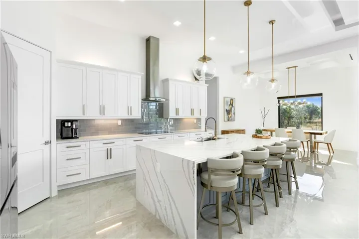 Kitchen featuring hanging light fixtures, wall chimney exhaust hood, an island with sink, tasteful backsplash, and white cabinetry