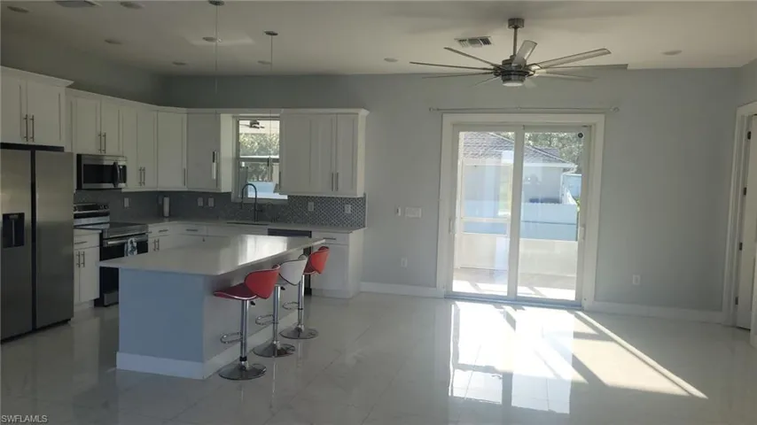 Open-concept kitchen and living area featuring white cabinetry, a central island with a light-colored countertop, stainless steel appliances, and a mosaic tile backsplash