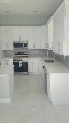 Modern kitchen featuring white shaker cabinetry, stainless steel appliances, a light-toned countertop, a gray mosaic tile backsplash, and large format floor tiling