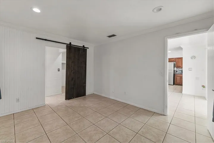 Empty room with a barn door, light tile patterned floors, baseboards, visible vents, and crown molding