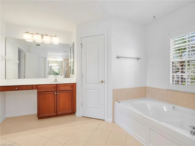 Bathroom featuring a built-in soaking tub with tiled surround, plantation shutters, and a vanity with a white countertop and wood-finish cabinetry