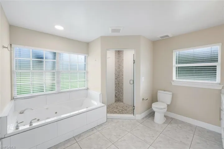 Spacious bathroom featuring a built-in jetted tub, a separate walk-in shower with glass enclosure, white tile flooring, and multiple windows