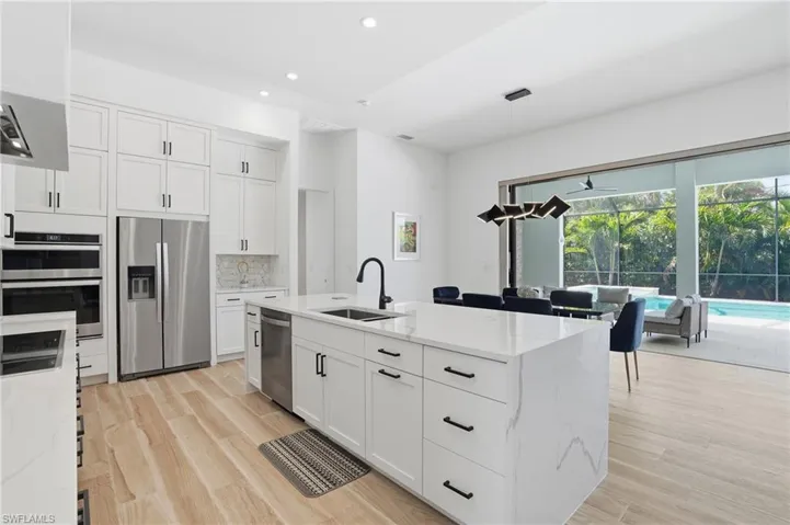 Kitchen with stainless steel appliances, hanging light fixtures, an island with sink, light stone counters, and light wood-style flooring