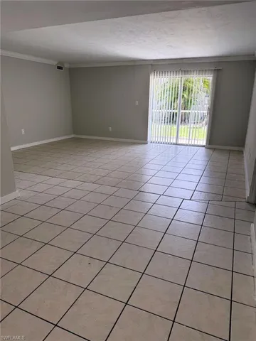 Spacious interior room featuring light-toned tile flooring with dark grout, neutral wall paint, and white crown molding