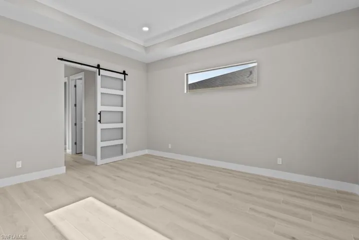 Masterbed room with a barn door, ornamental molding, light wood finished floors, baseboards, and a tray ceiling