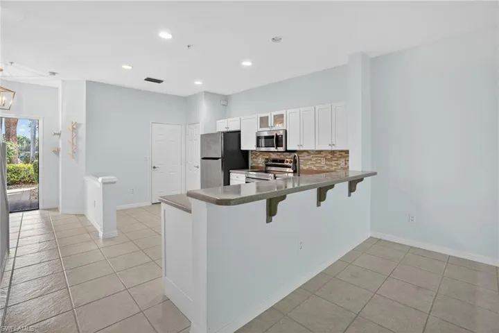 Open-concept kitchen and living area featuring tile flooring, white cabinetry, a breakfast bar with a stone-finish countertop, stainless steel appliances, and a mosaic tile backsplash