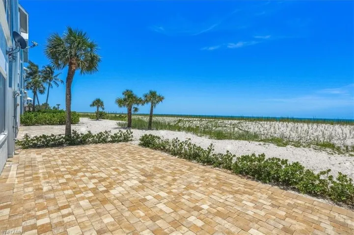Spacious paver patio with direct beach access, surrounded by sandy landscaping and mature palm trees