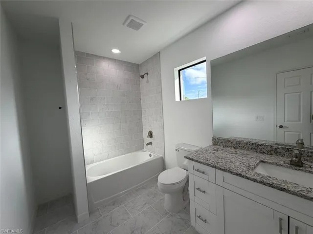 Bathroom featuring a vanity with stone-finish countertop, white cabinetry, integrated sink, bathtub with stone-tile surround, and stone-finish floor tile